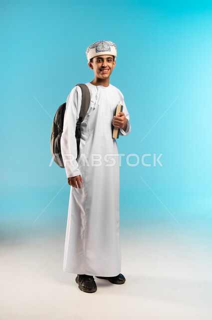 الاستعداد للذهاب إلى المدرسة، ايماءات تدل على السعادة و السرور، بورتريه لولد عربي خليجي عماني يرتدي الدشداشة و الكمه يحمل على ظهرة حقيبة مدرسية وبيده كتاب ، بدء العام الدراسي الجديد في سلطنة عمان، صورة الجسم بالطول الكامل, خلفية زرقاء .