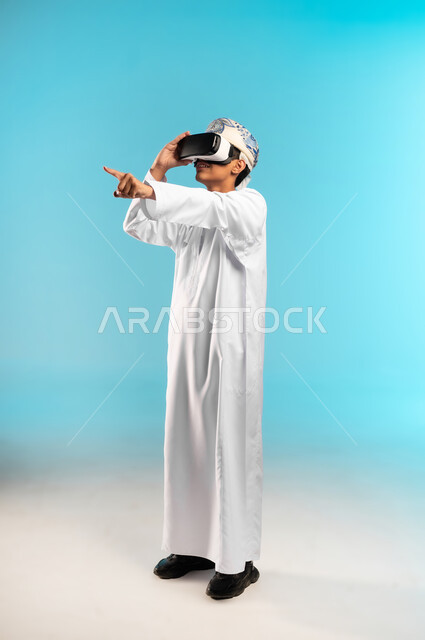 النظر الى الافلام الواقعيه ، استخدام جهاز تقني حديث ومتطور، محاكاة العالم الخيالي، بورتريه لولد عربي خليجي عماني يرتدي الدشداشه والكمه ينظر من خلال نظارات ثلاثية الأبعاد (الفي ار) بإيماءات السرور, المتعة والتسلية بالمشاهدة عبر نظارة الواقع الافتراضي، الجسم بالطول الكامل، خلفية زرقاء .