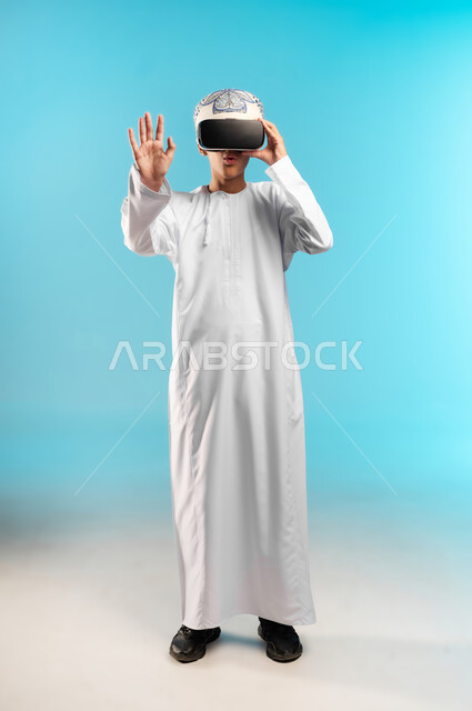 Using a modern and advanced technical device, simulating the imaginary world, a portrait of an Arab Gulf Omani boy wearing a dishdasha and a cloak, looking through 3D glasses (VR) with gestures of pleasure, enjoyment and entertainment by watching through virtual reality glasses, full-length body, blue background.