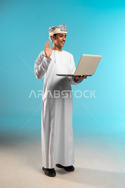 اجراء بث مباشر والقاء التحية ، التقنيه والتكنولوجيا الحديثه، التعليم الالكتروني ، بورتريه لصبي عربي خليجي عماني يرتدي الدشداشة والكمة يشير بيده نحو جهاز الحاسوب المحمول، صورة لكامل الجسم، خلفية زرقاء .