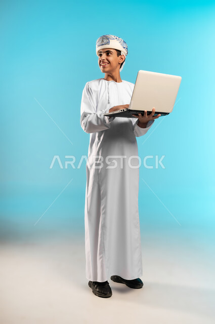 مراجعة الدروس باستخدام الحاسوب المحمول، اتباع طرق تعليمية جديدة، مدارس وأكاديميات متطوره، التقنيه والتكنولوجيا الحديثه، التعليم الالكتروني ، بورتريه لصبي عربي خليجي عماني يرتدي الدشداشة والكمة يشير باصبعه لمكان ما، صورة لكامل الجسم، خلفية زرقاء .