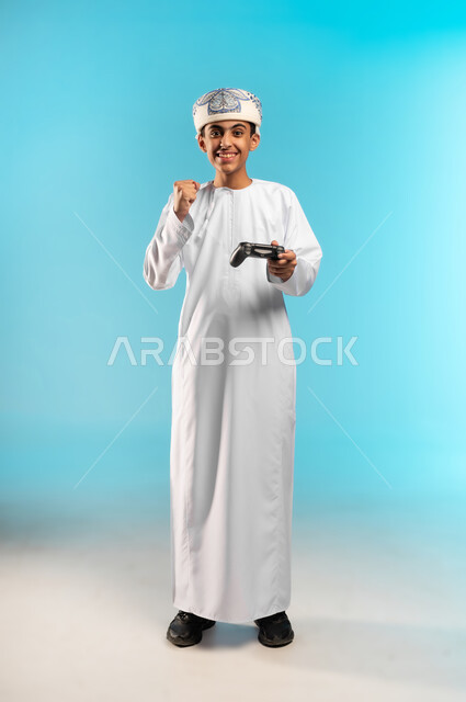 رفع قبضة اليد بتعابير الفوز والحماس ، التركيز والإندماج ، ألعاب الفيديو الترفيهية ، بورتريه لولد عربي عماني خليجي يرتدى الدشداشة والكمة  ، يحمل بيده يد التحكم عن بعد ، تعابير وجه تدل على السعادة و الحماس ، تقنيات وألعاب حديثة ، قضاء أوقات ممتعة ، خلفية زرقاء.