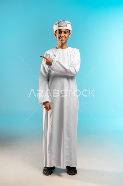 التوضيح والاشارة إلى شيء ما، بورتريه لولد عربي خليجي عماني يرتدي العمامة والدشداشة ويشير بإصبعه السبابة إلى مكان ما، تحديد موقع معين بايماءات السعادة والسرور، إشارات وحركات يدين تدل على هذا المكان (هنا)، صورة الجسم بالطول الكامل, خلفية زرقاء .