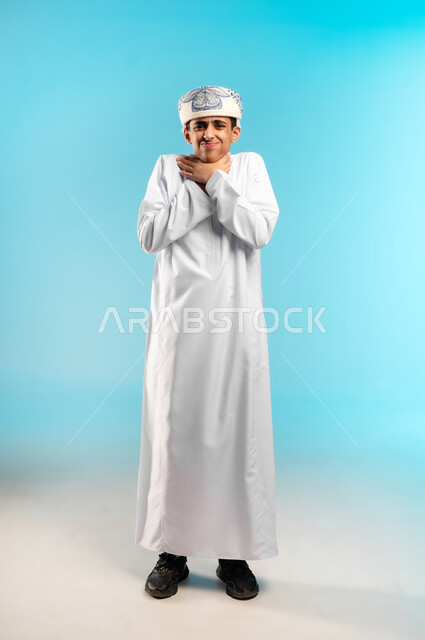 الاحساس بألم البلعوم والحاجة إلى العلاج والدواء, مفهوم الاهتمام بالصحة, الاصابة بنزلة برد والتهاب الحلق, بورتريه لولد عربي خليجي عماني يرتدي الدشداشه والكمه يضع يده على حلقه بايماءات الشعور بالم الحلق والاحتقان, خلفية زرقاء .