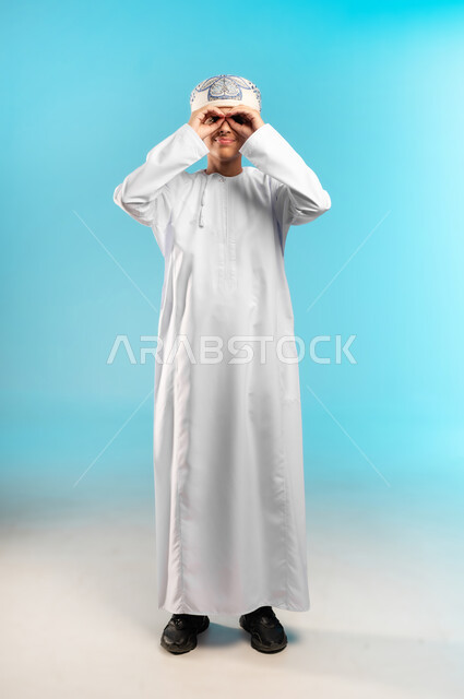 الوقوف امام الكاميرا وأداء حركات تمثيليه، بورتريه لولد عربي خليجي عماني يرتدي الدشداشة التقليدية والكمه العمانية يضع يديه على عينيه بشكل دائري بتمثيل حركه المنظار، التأمل بتحقيق الأحلام والطموحات ورؤية الخطط المستقبلية، خلفية زرقاء .