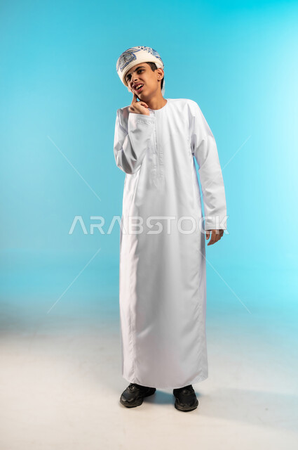 Focus, absent-mindedness and contemplation, finding a solution to a problem, contemplation and planning for the future, portrait of an Arab Gulf Omani boy wearing a dishdasha and a kummah, standing straight, placing his hand on his chin and looking at the camera with thinking gestures, full body image, blue background.
