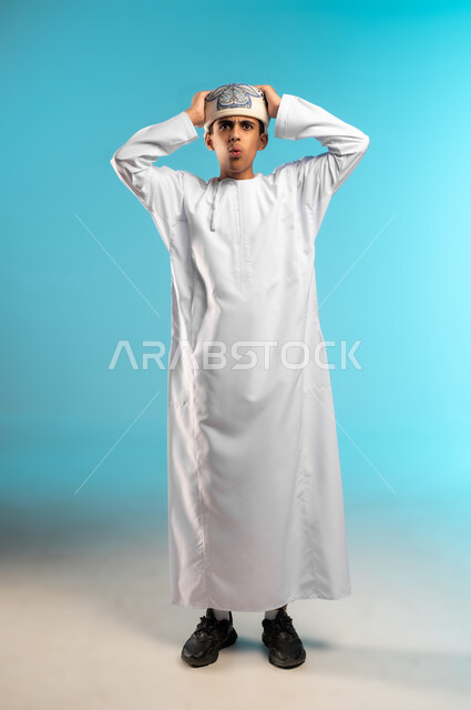 التفاجؤ من سماع خبر سيء، الشعور بالدهشة والصدمة والذهول, بورتريه لولد عربي خليجي عماني يرتدي الدشداشة والكمة ينظر الى الكاميرا يضع يديه على رأسه بإيماءات الخوف والذعر, صورة الجسم بالطول الكامل, خلفية زرقاء .