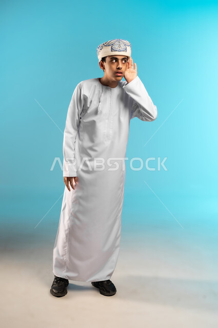 الانصات وحسن الاستماع, التركيز على الكلام و محاولة سماع الأصوات، حدوث ضعف في السمع, بورتريه لولد عربي خليجي عماني يرتدي الدشداشة والكمة يضع يده خلف اذنه بإيماءات التنصت, الجسم بالطول الكامل, خلفية زرقاء.