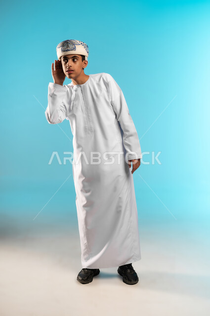 Focusing on speech and trying to hear sounds, listening and good listening, hearing loss, portrait of an Arab Gulf Omani boy wearing a dishdasha and a kummah, putting his hand behind his ear with eavesdropping gestures, full body, blue background.