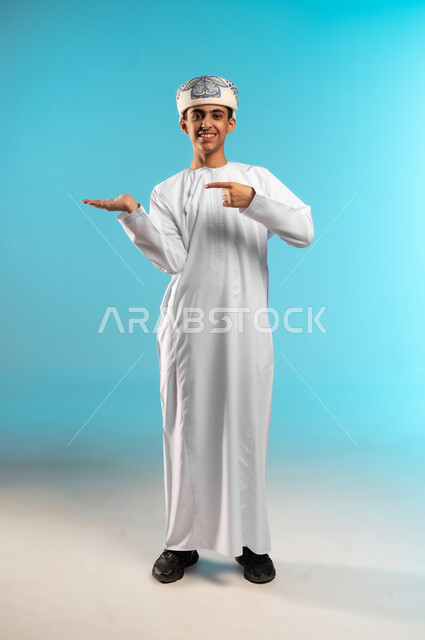 تحديد موقع أو هدف معين، الدلالة على مكان عنصر، بورتريه لولد عربي خليجي عماني مبتسم يرتدي الدشداشة والكمة يشير بيده إلى كف اليد الأخرى ينظر إلى الكاميرا بإيماءات السرور، حمل ورفع شيء ما، مفهوم الإشارة والتوجيه، الجسم بالطول الكامل، خلفية زرقاء .