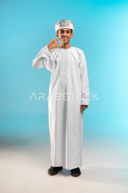 Pointing to the mouth with gestures of discomfort and pain, gum problems, requesting treatment and help, need to visit the dentist, portrait of an Arab Gulf Omani boy wearing a dishdasha and a kummah putting his hand over his mouth, full body image, blue background.