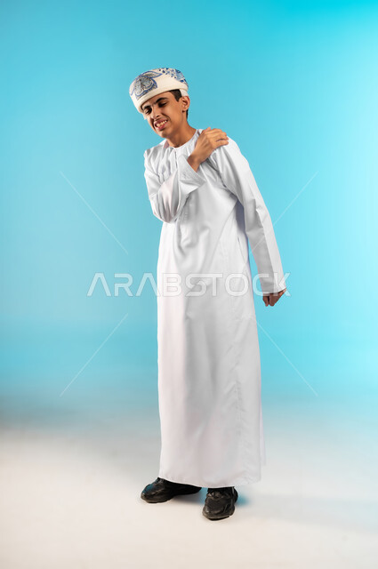 التهاب العضلات والمفاصل، الإحساس بالتعب والإرهاق, إيماءات الألم في الأكتاف، بورتريه لولد عربي خليجي عماني يرتدي الدشداشة والكمة يضع يده على كتفه بتعابير الوجع، الحاجه لوضع خطة علاجية, طلب المساعده للذهاب الى المستشفى، صورة الجسم بالطول الكامل، خلفية زرقاء .
