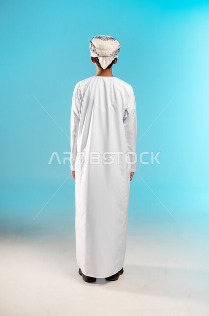 The concept of self-confidence, the concept of elegance and interest in external appearance, a portrait from the back of an Arab Gulf Omani boy wearing a dishdasha and a sleeve looking at something with smiling gestures, a full-length body image, blue background.