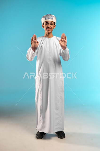 The order to stop and not to go further, a request to stop doing something, refusal and not to submit, a prohibition against advancing and the importance of leaving a safe distance, a portrait of an Arab Gulf Omani boy wearing a kummah and a dishdasha, raising his hand up in gestures of anger and displeasure, full-length body, blue background.