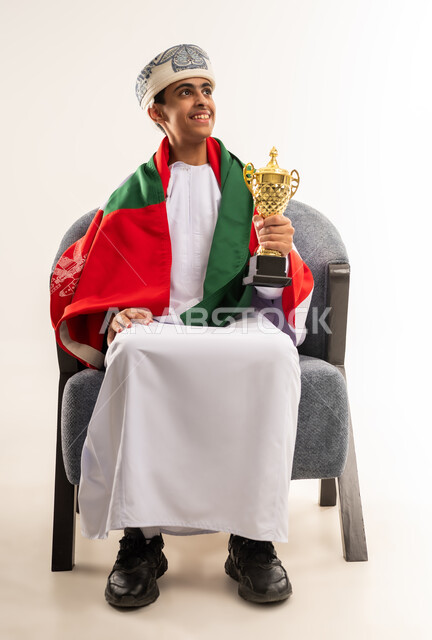 ارتداء اوشحة تشجيعية للفريق المفضل، الفرح والسرور لفوز المنتخب الوطني ، ايماءات الحماس والتشجيع، تشجيع الفريق العماني، بورتريه لولد عربي خليجي عماني يرتدي الدشداشة والكمة ووشاح ينظر الى الاعلى يحمل بيده كأس ، صورة الجسم بالطول الكامل, خلفية بيضاء .