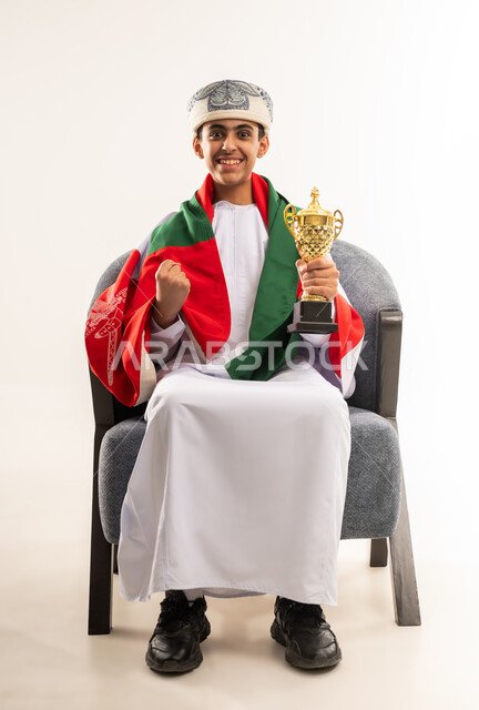 رفع قبضة اليد بتعابير الفوز، الفرح والسرور لفوز المنتخب الوطني ، ايماءات الحماس والتشجيع، تشجيع الفريق العماني، بورتريه لولد عربي خليجي عماني يرتدي الدشداشة والكمة ووشاح ينظر الى الاعلى يحمل بيده كأس ، صورة الجسم بالطول الكامل, خلفية بيضاء .