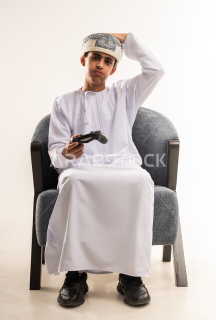 ايماءات وجه تدل على العبوس والإحباط والحزن  ، التركيز والإندماج ، ألعاب الفيديو الترفيهية ، بورتريه لولد عربي عماني خليجي يرتدى الدشداشة والكمة ، يجلس على كرسي ، يحمل بيده يد التحكم عن بعد ، تعابير وجه تدل على السعادة و الحماس ، تقنيات وألعاب حديثة  ، قضاء أوقات ممتعة ، خلفية بيضاء .