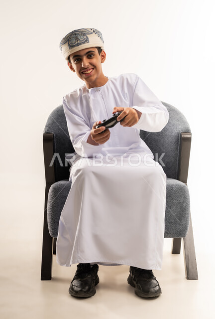 Focus and integration, entertainment video games, portrait of an Omani Gulf Arab boy wearing a dishdasha and a kummah, sitting on a chair, holding a remote control, facial expressions indicating happiness and enthusiasm, modern technology and games, having a good time, white background.
