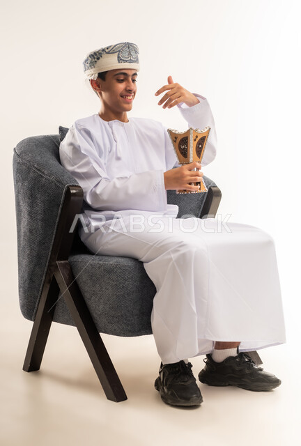 التطيب بالعطور والبخور, استخدام خشب العود بالأعياد والمناسبات, بورتريه لولد عربي خليجي عماني يرتدي الدشداشه والكمه يحمل بيده المبخرة ويقوم بالتعطر, الأناقة والاهتمام بالمظهر الخارجي, العادات والتقاليد العمانيه, خلفيه بيضاء .
