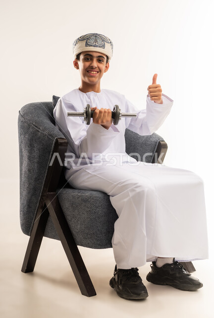 بناء عضلات الجسم وتقويتها، رفع الأوزان الحديدية الثقيلة، نشاط بدني للمحافظة على اللياقة الجسدية، بورتريه لولد عربي خليجي عماني يرتدي الدشداشة والكمة يجلس على كرسي مريح يحمل بيده الأثقال، خلفية بيج .