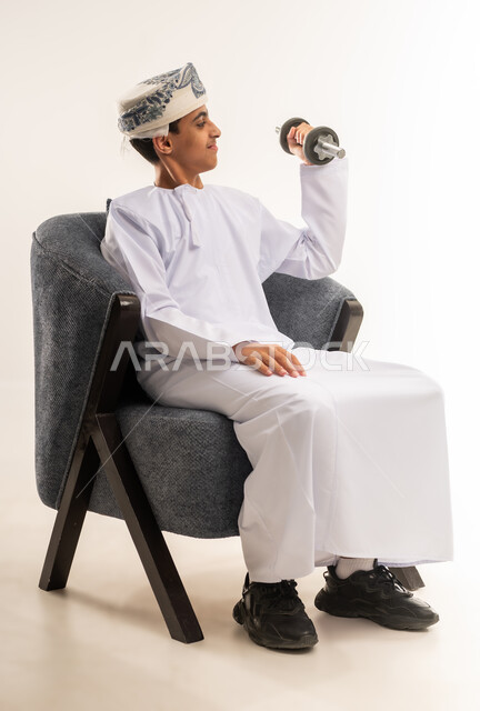 رفع الأوزان الحديدية الثقيلة، نشاط بدني للمحافظة على اللياقة الجسدية، بناء عضلات الجسم وتقويتها، بورتريه لولد عربي خليجي عماني يرتدي الدشداشة والكمة يجلس على كرسي مريح يحمل بيده الأثقال، خلفية بيج .