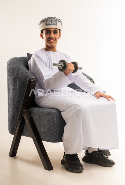 Physical activity to maintain physical fitness, lifting heavy iron weights, building and strengthening the body muscles, portrait of an Arab Gulf Omani boy wearing a dishdasha and a kumrah sitting on a comfortable chair holding weights in his hand, beige background.
