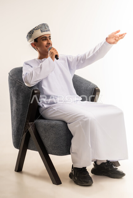 تقديم العروض الصوتية، تعابير الاندماج والتفاعل مع الموسيقى ، بورتريه لولد عربي خليجي عماني مبتسم يرتدي الكمة و الدشداشة يمسك بيده المايكروفون يقوم بالغناء ينظر الى الكاميرا بإيماءات الفرح والسرور، تسجيل مقطع صوتي غنائي، خلفية بيج .