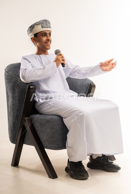 تعابير الاندماج والتفاعل مع الموسيقا، تسجيل مقطع صوتي غنائي، بورتريه لولد عربي خليجي عماني مبتسم يرتدي الكمة و الدشداشة يمسك بيده المايكروفون يقوم بالغناء ينظر الى الكاميرا بإيماءات الفرح والسرور، تقديم العروض الصوتية، خلفية بيج .