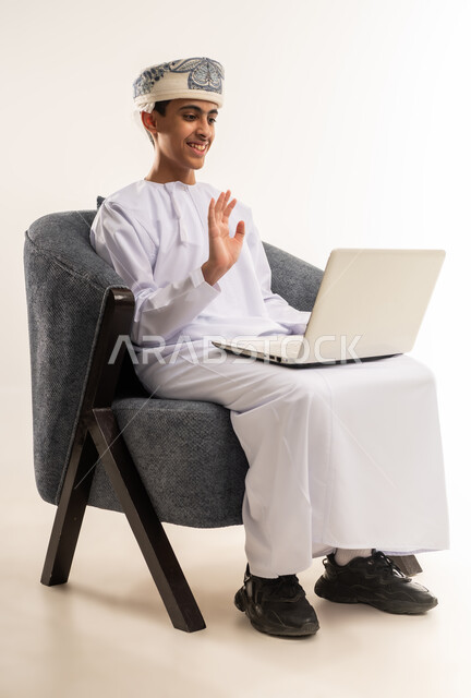 القاء التحية عبر مكالمات الفيديو، استخدام الاجهزة التقنية الحديثة، مفهوم التعليم الإلكتروني، متابعة الحصص وحل الواجبات عبر الانترنت، بورتريه لولد عربي خليجي عماني يرتدي الدشداشة والكمة يقوم باجراء بث مباشر، المذاكرة والدراسة عن بعد، خلفية بيج .