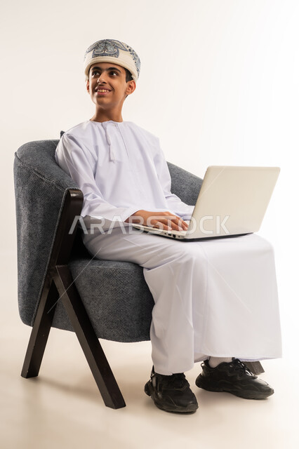 استخدام الاجهزة التقنية الحديثة، مفهوم التعليم الإلكتروني، اجراء مكالمات الفيديو،  متابعة الحصص وحل الواجبات عبر الانترنت، بورتريه لولد عربي خليجي عماني يرتدي الدشداشة والكمة يقوم بمراجعة دروسه، المذاكرة والدراسة عن بعد، خلفية بيج .
