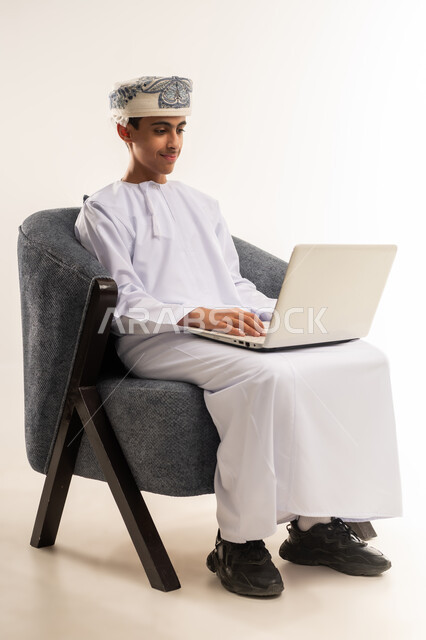The concept of e-learning, the use of modern technical devices, making video calls, following classes and doing homework online, a portrait of an Arab Gulf Omani boy wearing a dishdasha and a kuma reviewing his lessons, studying and studying remotely, beige background.