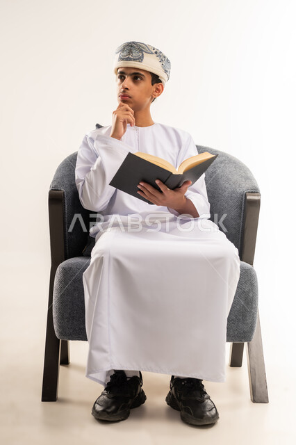 Love of reading, spending free time educating and learning, developing reading skills, interest in browsing useful books, portrait of an Arab Gulf Omani boy wearing a dishdasha and a kummah sitting on a comfortable chair holding a book in his hand, beige background.