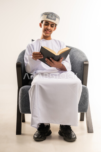 النظر إلى الكتاب بابتسامة, حب المطالعة والقراءة, قضاء أوقات الفراغ  بالتثقيف والتعلم, تنمية مهارات القراءة, الاهتمام بتصفح الكتب المفيدة, بورتريه لولد عربي خليجي عماني يرتدي الدشداشة والكمة يجلس على كرسي مريح ويمسك بيده الكتاب, خلفية بيج .