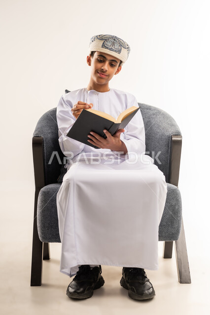 مفهوم الجد والاجتهاد ، حب المطالعة والقراءة, قضاء أوقات الفراغ  بالتثقيف والتعلم, تنمية مهارات القراءة, الاهتمام بتصفح الكتب المفيدة, بورتريه لولد عربي خليجي عماني يرتدي الدشداشة والكمة يجلس على كرسي مريح ويمسك بيده الكتاب وقلم , خلفية بيج .