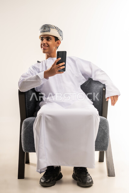 تبادل الأحاديث مع الأهل والأصدقاء، استخدام التكنولوجيا والأجهزة التقنية الحديثة المتطورة، ايماءات السعادة والفرح، بورتريه لولد عربي خليجي عماني يرتدي الدشداشة والكمة مبتسم يمسك بيده الهاتف المحمول، الجسم بالطول الكامل, خلفية بيج .