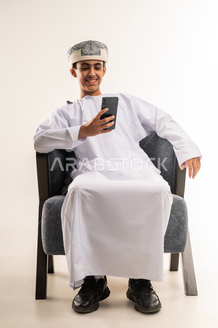 استخدام التكنولوجيا والأجهزة التقنية الحديثة المتطورة، تبادل الأحاديث مع الأهل والأصدقاء، ايماءات السعادة والفرح، بورتريه لولد عربي خليجي عماني يرتدي الدشداشة والكمة مبتسم بيده الهاتف المحمول، الجسم بالطول الكامل, خلفية بيج .