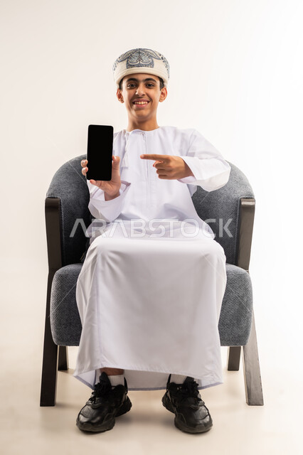 عرض شاشة سوداء فارغة عبر الهاتف المحمول, استخدام جهاز تقني حديث ومتطور, بورتريه لولد عربي خليجي عماني يرتدي الدشداشة والكمة يجلس على كرسي يقوم يمسك بيده الجوال, الجسم بالطول الكامل, خلفية بيج .
