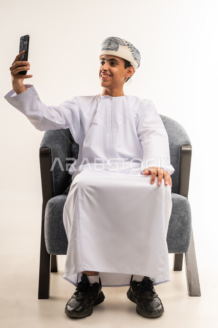 توثيق اللحظات السعيدة عبر الهاتف المحمول, استخدام جهاز تقني حديث ومتطور, بورتريه لولد عربي خليجي عماني يرتدي الدشداشة والكمة يجلس على كرسي يقوم بالتقاط صورة السيلفي التذكارية عبر الجوال, الجسم بالطول الكامل, خلفية بيج .