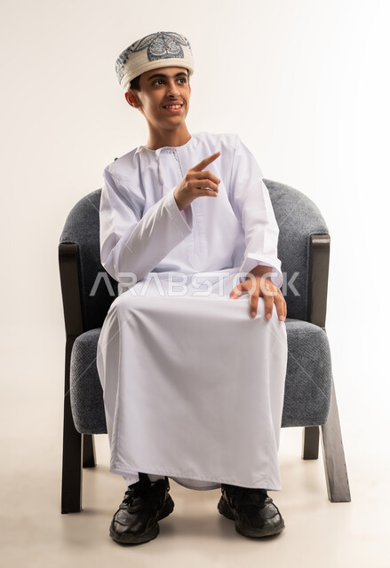 الاشارة جانباً الى شئ ما ، بورتريه لولد عربي خليجي عماني مبتسم يرتدي الدشداشة والكمه ينظر الى الكاميرا يرفع يده للاعلى يشير بأصبع السبابة الى هدف محدد، مفهوم النقر والضغط والتفاعل مع العناصر الهولوجرامية، الجسم بالطول الكامل، خلفية بيج .