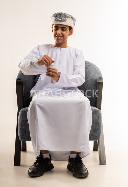 اهمية الإصرار والمثابرة ، مفهوم القلق أو التوتر الخفيف، بورتريه لولد عربي خليجي عماني يرتدي الدشداشه والكمه يجلس على كرسي مريح يقوم محاولة فتح علبة فارغة ، التفاعل مع الاشياء فى الحياة اليومية ، الجسم بالطول الكامل، خلفية بيضاء .