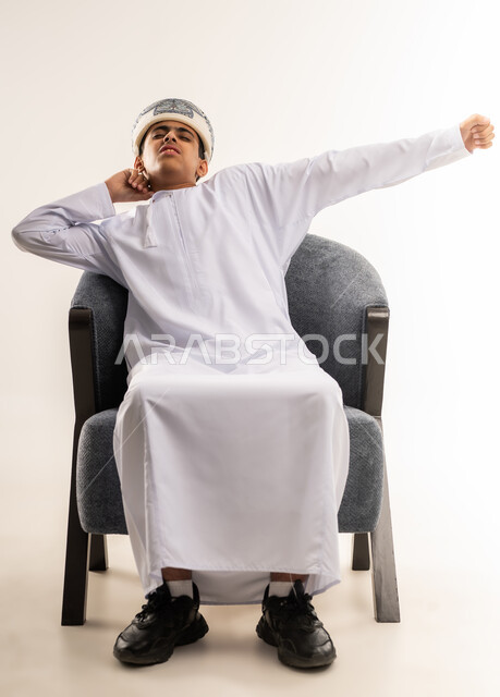 The need to sleep and feeling tired and lethargic, the feeling of tiredness and exhaustion and the need to take some rest, a portrait of an Arab Gulf Omani boy wearing a kummah and a dishdasha sitting on a comfortable chair with his hands raised in a sleepy gesture, beige background.