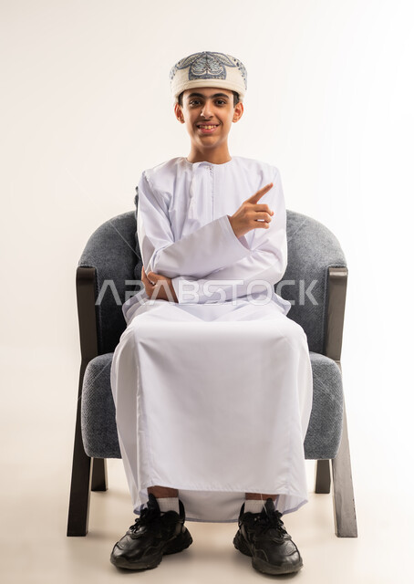 The concept of clicking, pressing and interacting with holographic elements, determining a specific location, a portrait of a smiling Omani Gulf Arab boy wearing a dishdasha and a sleeve sitting on a chair looking at the camera, raising his hand up and pointing with his index finger to a specific target, full body, beige background.