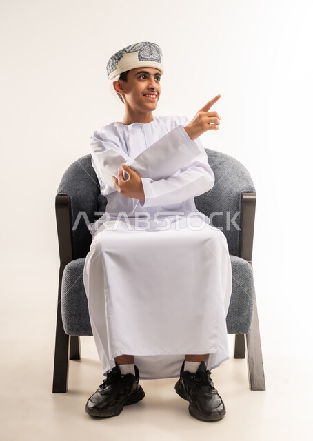 تحديد موقع معين، بورتريه لولد عربي خليجي عماني مبتسم يرتدي الدشداشة والكمه يجلس على كرسى ينظر الى الكاميرا ، يرفع يده للاعلى يشير بأصبع السبابة الى هدف محدد، مفهوم النقر والضغط والتفاعل مع العناصر الهولوجرامية، الجسم بالطول الكامل، خلفية بيج .