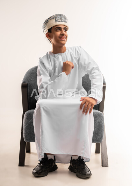 الشعور بالسعادة والقوة، مفهوم الانجاز وتحقيق الاهداف، بورتريه لولد عربي خليجي عماني مبتسم يرتدي الدشداشة و الكمه ينظر الى الكاميرا يرفع قبضة يده للاعلى بإيماءات الفرح والسرور, الانتصار والفوز والنجاح، الجسم بالطول الكامل، خلفية بيج .