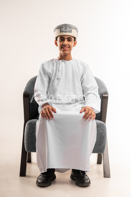Looking at the camera with gestures of joy, the concept of elegance and interest in outward appearance, sitting upright and confident, a portrait of a smiling Omani Gulf Arab boy wearing a dishdasha and a kummah sitting on a comfortable chair with expressions of happiness and pleasure, beige background.