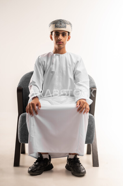 ايماءات الجدية والتركيز ، مفهوم الاناقه والاهتمام بالمظهر الخارجي، الجلوس باستقامة وثقه بالنفس، بورتريه لولد عربي خليجي عماني مبتسم يرتدي الدشداشة والكمة يجلس على كرسي مريح  ينظر الى الكاميرا ، خلفية بيج .
