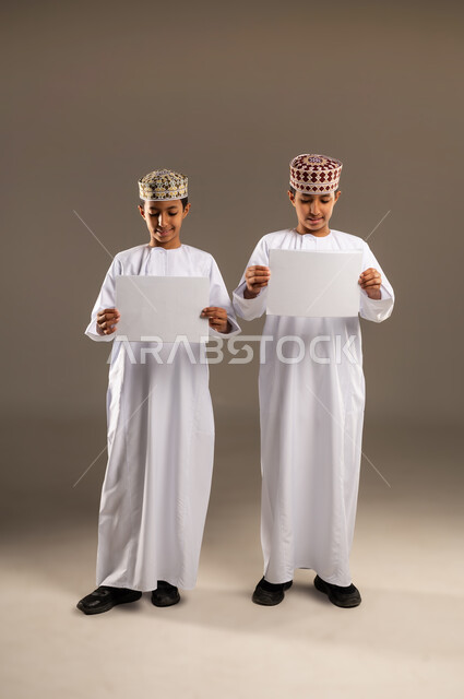 التعاون بين الأخوة في إنجاز مهام العمل, تبادل الخبرات والمهارات, مناقشة الخطط والمشاريع, بورتريه لولدان توأم عربيين خليجيين عمانيين يرتديان الدشداشة والكمة يمسكان بأيديهم الأوراق البيضاء, الجسم بالطول الكامل، خلفية بيج .