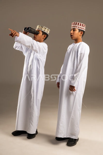 Planning for goals and ambitions, love of adventure and exploration, portrait of two Omani Gulf Arab boys wearing dishdasha and kummah, one of them looking through binoculars (telescope) and pointing at something with gestures of joy and pleasure, seeing distant things through binoculars, full body image, beige background.