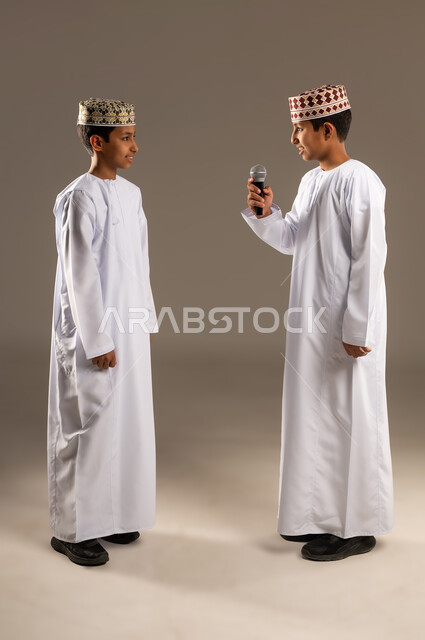 تسجيل مقطع صوتي غنائي، تقديم عرض فني وفقرة ترفيهية، بورتريه لولدان عربيان خليجيان عمانيان يرتديان الدشداشة والكمة احدهما يمسك مكبر الصوت ، اكتشاف موهبة الغناء منذ الصغر,،جهاز تقني حديث، الجسم بالطول الكامل، خلفية بيج .