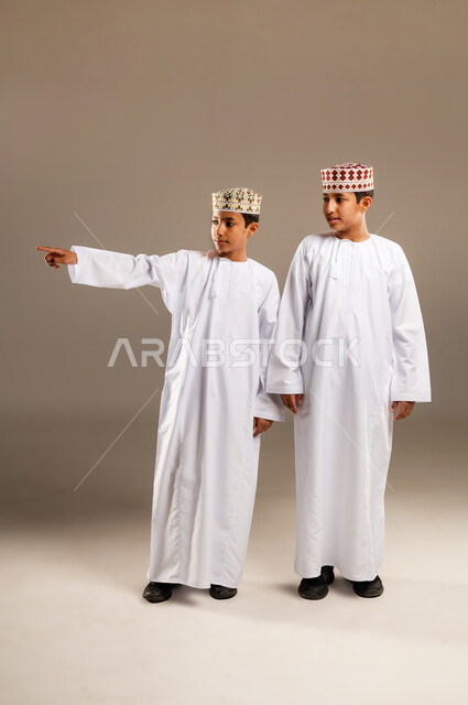 حركات تدل على هذا المكان (هنا), تعابير يدين تشير الى موقع معين, مفهوم لغة الجسد والإشارات، بورتريه لولدان توأم عربيان خليجيان عمانيان مبتسمان يرتديان الدشداشة والكمة ويشيران بأصابع السبابة إلى شيء ما بإيماءات السرور، خلفية بيج.